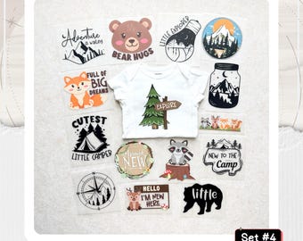 May include: A white baby bodysuit featuring a green pine tree graphic with a sign reading Explore, surrounded by various iron-on transfers including a bear face, a fox, a tent, a compass, a raccoon on a stump, and text like Adventure is waiting.