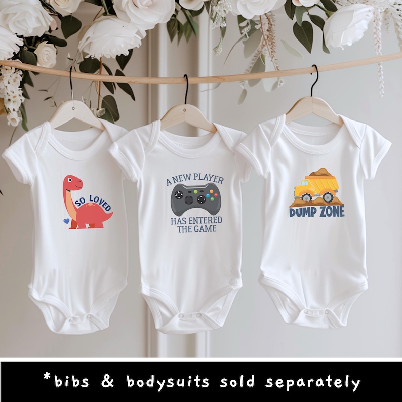 May include: Three white short-sleeve bodysuits hanging on wooden hangers. The first features a red dinosaur with the text SO LOVED. The second shows a black game controller with the text A NEW PLAYER HAS ENTERED THE GAME. The third displays a yellow dump truck with the text DUMP ZONE.