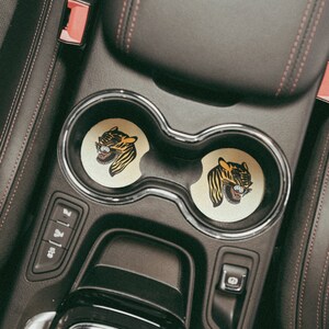Puede incluir: Dos portavasos negros con un dise&ntilde;o de cara de tigre en cada posavasos. Los posavasos son amarillos y negros con un contorno blanco. Los portavasos est&aacute;n en un coche.