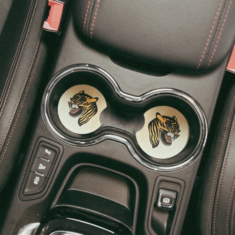 Puede incluir: Dos portavasos negros con un dise&ntilde;o de cara de tigre en cada posavasos. Los posavasos son amarillos y negros con un contorno blanco. Los portavasos est&aacute;n en un coche.