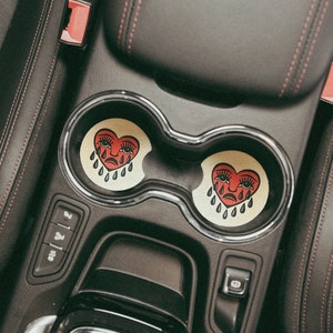 Puede incluir: Dos posavasos de coche en forma de coraz&oacute;n rojo con un dise&ntilde;o de cara llorando. Los posavasos est&aacute;n en los portavasos de un coche.
