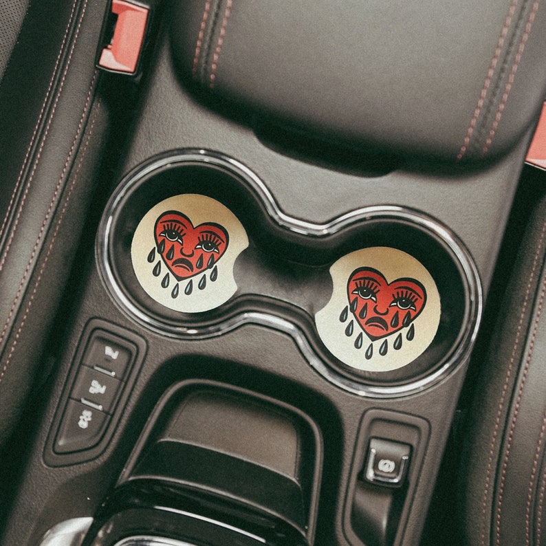 Puede incluir: Dos posavasos de coche en forma de coraz&oacute;n rojo con un dise&ntilde;o de cara llorando. Los posavasos est&aacute;n en los portavasos de un coche.