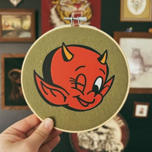 May include: A red and black cartoon devil with horns and a mischievous grin is embroidered on a green felt circle. The circle is surrounded by a brown wooden frame.
