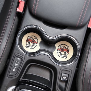 Puede incluir: Dos posavasos de coche en forma de calavera en blanco y negro con la palabra "SPEED" y gafas rojas en la calavera. Los posavasos est&aacute;n en el portavasos de un coche.