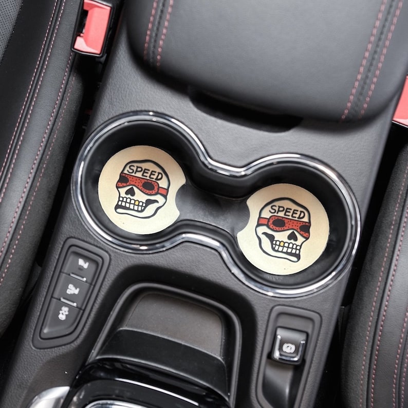 Puede incluir: Dos posavasos de coche en forma de calavera en blanco y negro con la palabra "SPEED" y gafas rojas en la calavera. Los posavasos est&aacute;n en el portavasos de un coche.