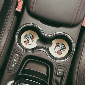 Puede incluir: Dos posavasos de coche beige con un gr&aacute;fico de una mano sosteniendo un coraz&oacute;n con una cadena alrededor de la mu&ntilde;eca. Los posavasos est&aacute;n en los portavasos de un coche.