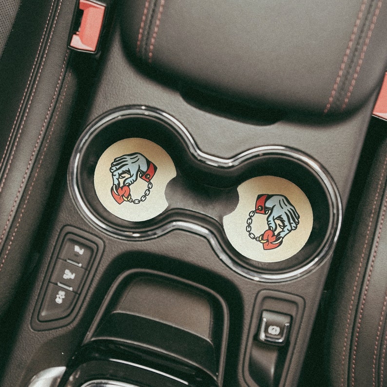 Puede incluir: Dos posavasos de coche beige con un gr&aacute;fico de una mano sosteniendo un coraz&oacute;n con una cadena alrededor de la mu&ntilde;eca. Los posavasos est&aacute;n en los portavasos de un coche.
