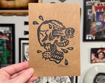 Puede incluir: Un grabado en linóleo en blanco y negro sobre cartulina marrón que representa una calavera con un ojo estilizado y lágrimas. La calavera tiene sombreado detallado y está rodeada de gotas de agua estilizadas. La obra de arte está sostenida por una mano.