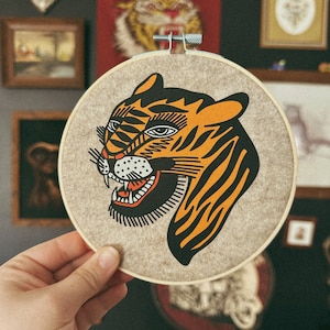 May include: A beige circular embroidery hoop with a black and orange tiger face design. The tiger's mouth is open and its eyes are black. The tiger's stripes are black and orange.