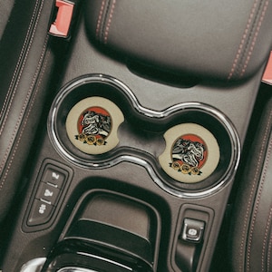 Puede incluir: Dos posavasos redondos de color marr&oacute;n con una ilustraci&oacute;n en blanco y negro de tres cabezas de caballo sobre un fondo rojo y flores amarillas alrededor de las cabezas de los caballos. Los posavasos est&aacute;n en un portavasos de coche.