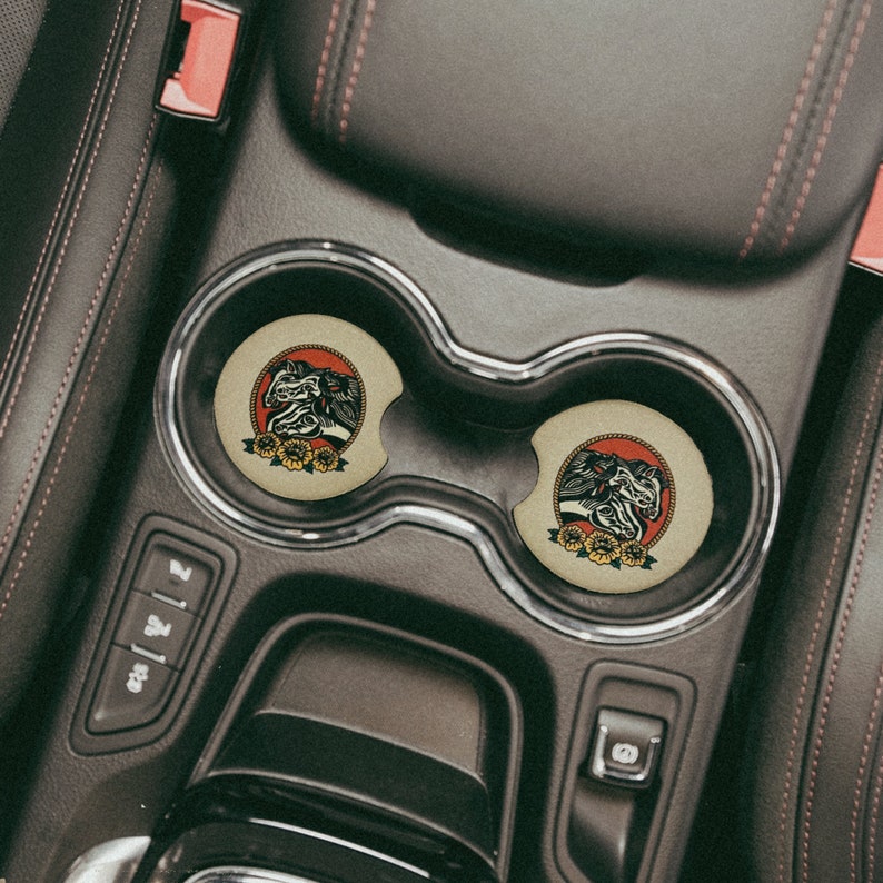 Puede incluir: Dos posavasos redondos de color marr&oacute;n con una ilustraci&oacute;n en blanco y negro de tres cabezas de caballo sobre un fondo rojo y flores amarillas alrededor de las cabezas de los caballos. Los posavasos est&aacute;n en un portavasos de coche.