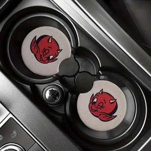 May include: Two gray car cup holders with red and yellow cartoon devil designs. The devil has horns, a red body, and a yellow face with a wink.