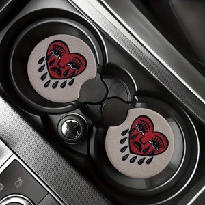 May include: Two gray car cup holders with red heart-shaped coasters. Each coaster features a black outline of a crying heart with eyelashes and black teardrop shapes.