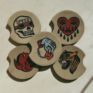 Puede incluir: Conjunto de cinco posavasos redondos con diferentes dise&ntilde;os. Los posavasos son beige con contornos negros y presentan diversas ilustraciones. Un posavasos tiene una calavera con la palabra "SPEED" encima. Otro posavasos tiene un coraz&oacute;n con una cara llorando y l&aacute;grimas. Un tercer posavasos tiene una mano sosteniendo un coraz&oacute;n con una cadena. Un cuarto posavasos tiene una cara de tigre. El quinto posavasos tiene una cara de diablo.
