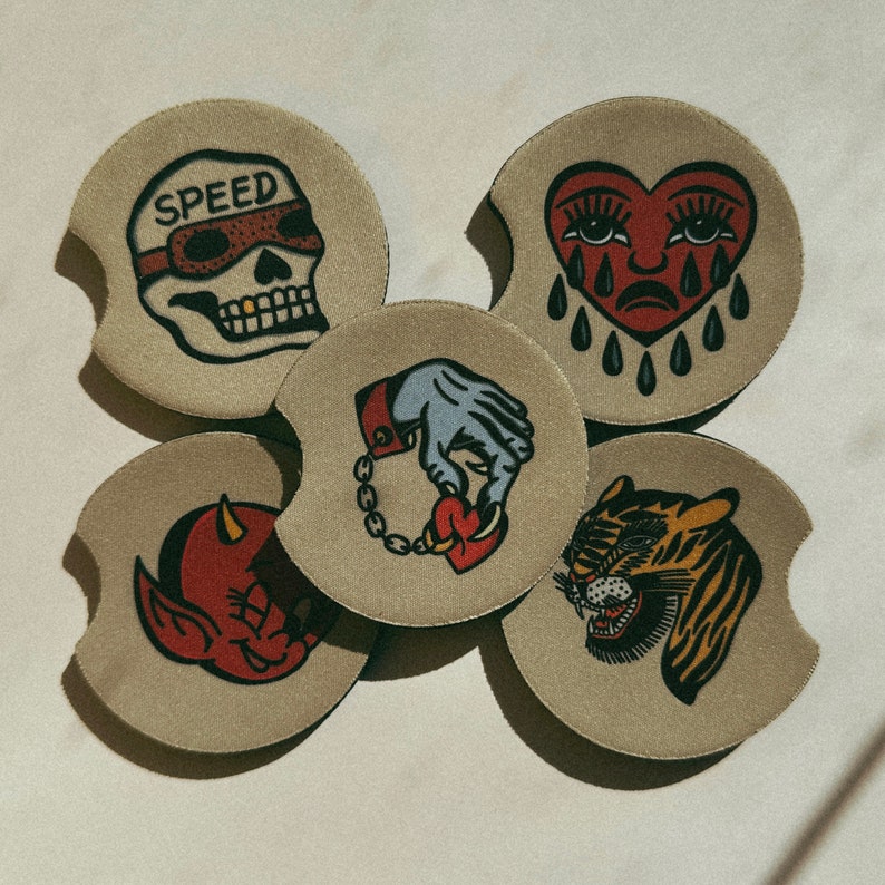 Puede incluir: Conjunto de cinco posavasos redondos con diferentes dise&ntilde;os. Los posavasos son beige con contornos negros y presentan diversas ilustraciones. Un posavasos tiene una calavera con la palabra "SPEED" encima. Otro posavasos tiene un coraz&oacute;n con una cara llorando y l&aacute;grimas. Un tercer posavasos tiene una mano sosteniendo un coraz&oacute;n con una cadena. Un cuarto posavasos tiene una cara de tigre. El quinto posavasos tiene una cara de diablo.