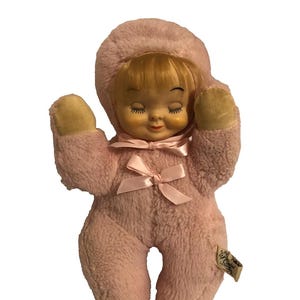 May include: A vintage pink plush doll with a hooded head, closed eyes, and a small pink bow. The doll has blonde hair and beige mittens. A tag is attached to the doll's leg.