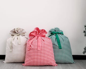 May include: Three large fabric gift bags in a gingham pattern. The bags are beige, red, and green, each with a matching ribbon tie at the top. The bags are standing upright on a wooden floor.