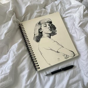 以下が含まれることがあります： 閉じた目の人物の白黒ポートレートが描かれた、スパイラル綴じのスケッチブック。 黒いペンが白いシーツの上にあるスケッチブックの隣に置かれています。 この絵はミニマリストスタイルです。