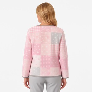 Pu&ograve; includere: Un top rosa a maniche lunghe con patchwork e rifiniture grigie intorno allo scollo e ai polsini. Il top presenta vari quadrati rosa e grigi a fantasia, tra cui stelle e quadretti. La modella indossa pantaloni grigi.