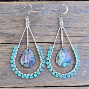 May include: A pair of teardrop-shaped earrings featuring silver-toned wire frames wrapped with small turquoise beads. Each earring contains a central iridescent abalone shell pendant suspended from a coiled silver wire post and a hook ear wire.