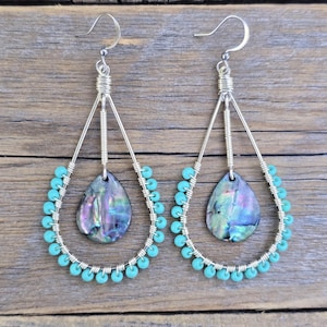 May include: A pair of teardrop-shaped earrings featuring silver-toned wire frames wrapped with small turquoise beads. Each earring contains a central, iridescent abalone shell pendant suspended from a silver wire coil and hook.