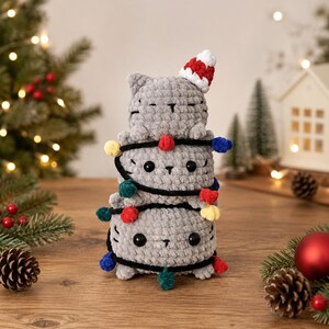May include: A stack of three gray crocheted cat plushies, with the top cat wearing a small red and white striped hat. A black string with colorful pom-poms in red, yellow, blue, and green is wrapped around the three stacked cat figures.