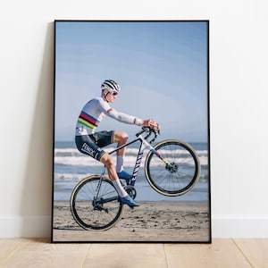 Può includere: Un ciclista con una maglia bianca e arcobaleno esegue una ruota anteriore su una bicicletta nera e blu su una spiaggia sabbiosa. Il ciclista indossa un casco e occhiali da sole.