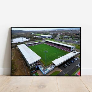 May include: Aerial view of a football stadium with a green pitch, white stands, and a red and white seating area. The stadium is surrounded by a car park and a lake.