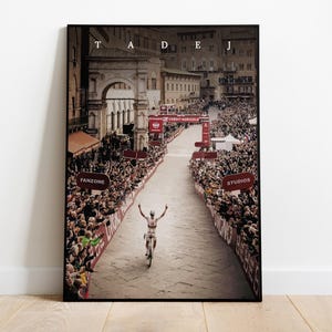 May include: A black and white photo of a cyclist raising their arms in victory as they cross the finish line of a race. The cyclist is wearing a white jersey with red and blue accents. The photo is taken from a high angle, looking down on the cyclist and the crowd of spectators lining the street. The street is lined with red and white banners and signs. The photo is titled "TADEJ" in white letters at the top.