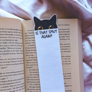 May include: A white bookmark with a black cat peeking over the top. The bookmark has the text "Is that smut again?" printed on it.