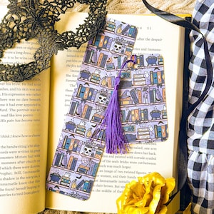 May include: Purple and black bookmark with a Halloween theme. The bookmark features a bookshelf with books, skulls, cats, potions, and other spooky items. The bookmark has a purple tassel.