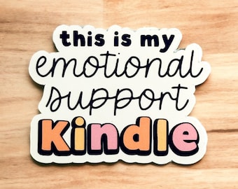 Puede incluir: Pegatina blanca con texto negro que dice "this is my emotional support Kindle" con la palabra "Kindle" en letras naranja y rosa.