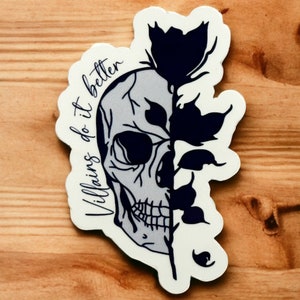 May include: A white sticker with a black and white skull and a black floral design. The text "Villains do it better" is written in a cursive font on the sticker.
