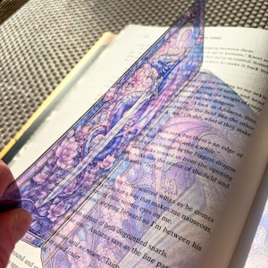 May include: A translucent rectangular bookmark featuring a stained-glass style illustration of a dragon coiled around a sword, rendered in shades of purple, blue, and pink. The design includes floral motifs and a crescent moon at the top edge.
