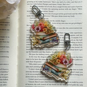 May include: A stack of colourful books with floral designs on the cover. The books are stacked on top of each other and are held together by a silver charm. The charm is shaped like a book and has a floral design on it. The books are surrounded by a white background.