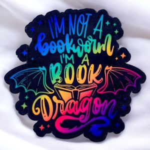 Op de afbeelding: Regenboogkleurige, iriserende sticker met de tekst "I'm not a bookworm I'm a book dragon" met een boek en drakenvleugels.