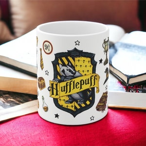 May include: White ceramic mug featuring the Hufflepuff crest from the Harry Potter series. The crest includes a badger, yellow and black stripes, and the word "Hufflepuff" on a banner. Additional icons and stars surround the crest.
