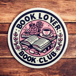 May include: A pink and white round sticker with a black border. The sticker features a book, a cup of coffee, and flowers. The text "BOOK LOVER BOOK CLUB" is written in a black circle around the image.