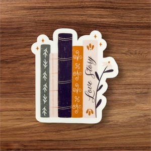 Può includere: Un adesivo con una pila di quattro libri con un motivo floreale e il testo "Love Story". I libri sono in tonalità di arancione, bianco e viola.