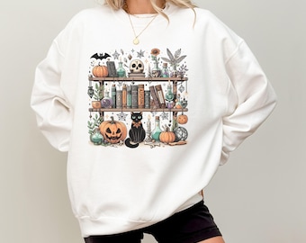Könnte beinhalten: Weißes Sweatshirt mit einem grafischen Druck eines Regals mit Büchern, Tränken, einer schwarzen Katze und Kürbissen. Das Regal ist mit Spinnweben, Kerzen und anderen Halloween-Artikeln dekoriert.