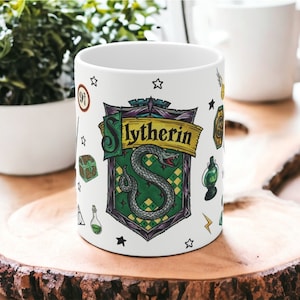 May include: White ceramic mug featuring the Slytherin house crest from the Harry Potter series. The crest includes a green snake on a shield, with the word "Slytherin" in gold. Additional small illustrations of magical items are scattered around the mug.