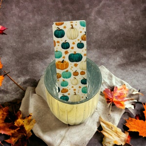 May include: A white and teal pumpkin patterned bookmark is placed inside a clear glass bowl with a gold rim. The bowl is sitting on a beige fabric surface with autumn leaves scattered around it.