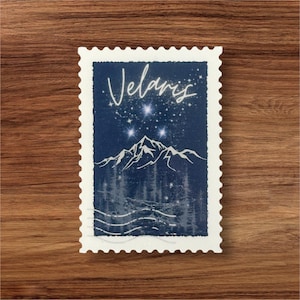 Può includere: Un francobollo blu e bianco con la parola "Velaris" scritta in corsivo bianco. Il francobollo presenta una catena montuosa con un cielo notturno stellato sopra.