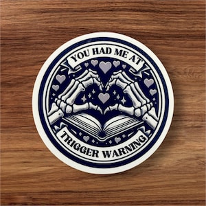 May include: A round sticker with a blue background and white text that reads "You had me at trigger warning". Two skeletal hands are forming a heart shape around an open book. The sticker has a white border.