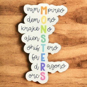 Peut inclure: Un autocollant blanc avec le mot "MONSTERS" écrit verticalement en couleurs arc-en-ciel. Chaque lettre est sur un autocollant séparé en forme de nuage. Les mots "vampire", "démon", "kraken", "alien", "shifter", "fée", "dragon" et "orc" sont écrits en cursive autour des lettres.