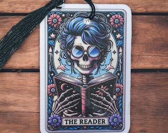 Può includere: Una carta bianca con un bordo nero e un'illustrazione colorata di uno scheletro con i capelli blu che legge un libro. Il testo "THE READER" è stampato nella parte inferiore della carta.