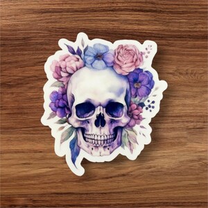Può includere: Un teschio bianco con una corona floreale viola e rosa. Il teschio ha un design dettagliato con denti e orbite visibili. I fiori sono dipinti in stile acquerello.
