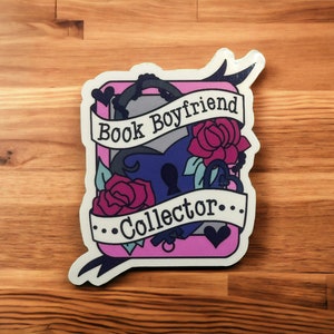 Può includere: Un adesivo con un bordo rosa e bianco con un lucchetto a forma di cuore, rose e le parole "Book Boyfriend Collector" in uno striscione. L'adesivo ha una combinazione di colori blu scuro e grigio.
