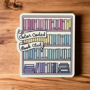 May include: A sticker featuring a cartoon bookshelf with rainbow coloured books. The bookshelf has the text "Color Coded Book Club" on it.