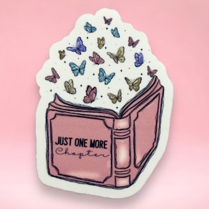 Può includere: Un libro rosa aperto con il testo "JUST ONE MORE CHAPTER" sulla copertina. Farfalle di vari colori volano fuori dal libro.